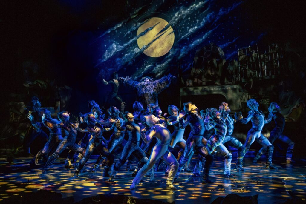 The Jellicle Cats dancing beneath a giant moon in the Cats the Musical junkyard set