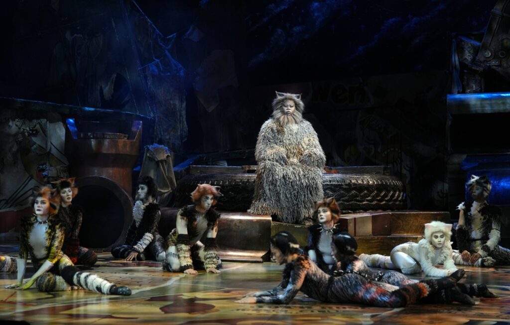 Old Deuteronomy seated on a tyre throne surrounded by the Jellicle Cats in Cats the Musical