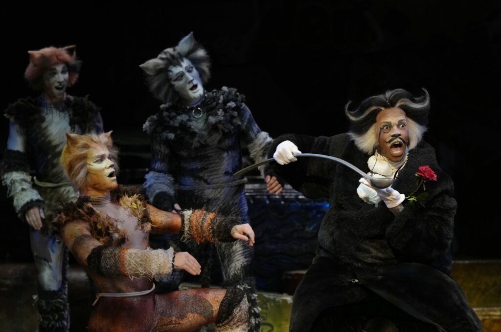 Bustopher Jones in a black coat and white spats holding a ladle with a rose in Cats the Musical